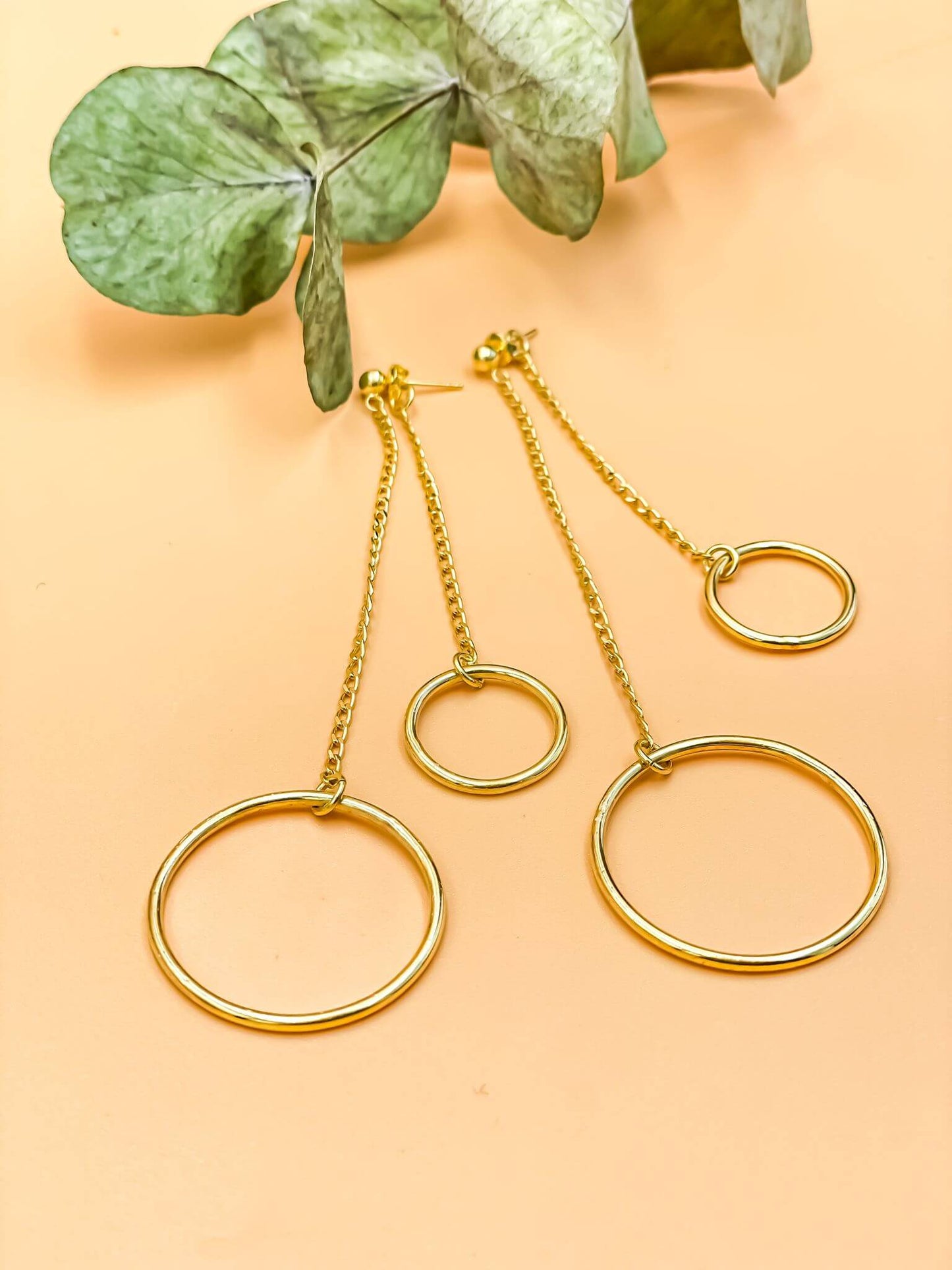 Harmony Swing Earrings in 18-carat gold plating with large and small hoops on a long chain, set on a sterling silver base alongside green leaves