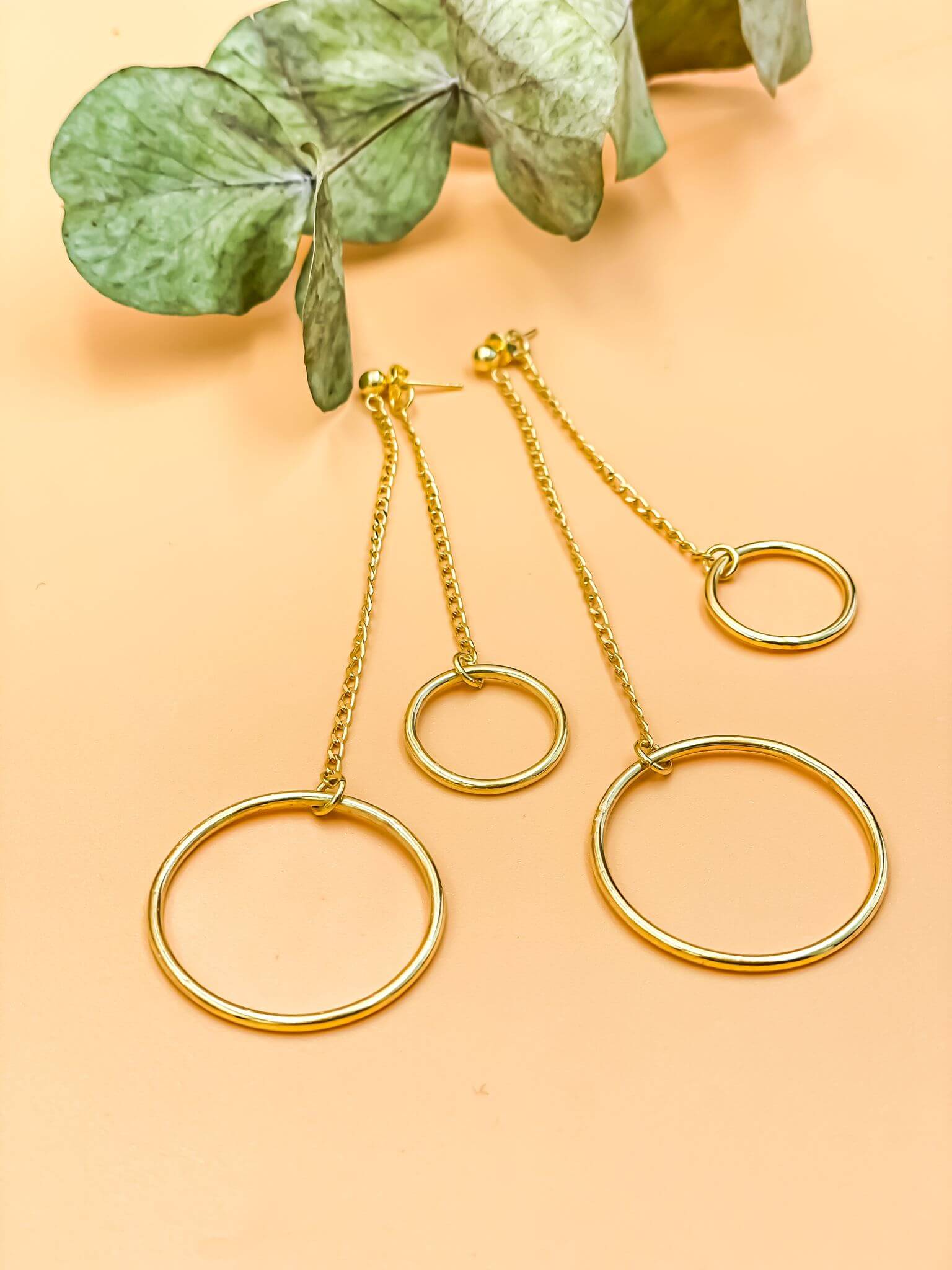 Harmony Swing Earrings in 18-carat gold plating with large and small hoops on a long chain, set on a sterling silver base alongside green leaves